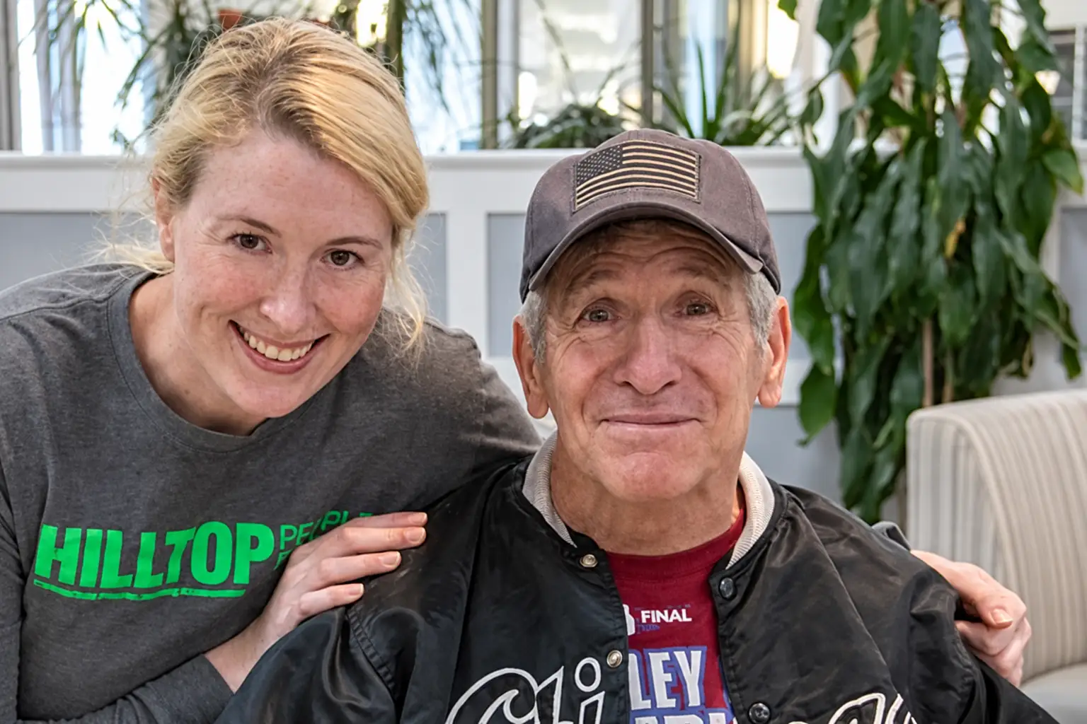 A woman wearing a Hilltop shirt rests her hand on an older man’s shoulder as they smile together.